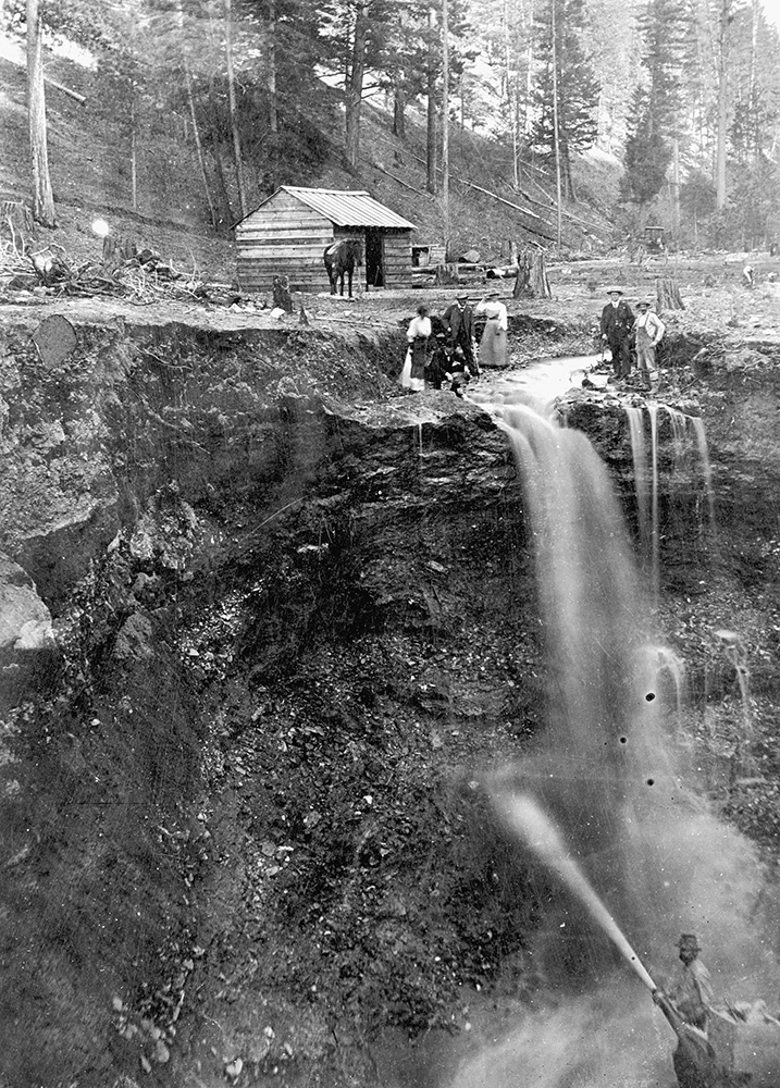 Several people stand at the opening to a deep hole looking down at a man with a hose shooting water at the side of the hole.